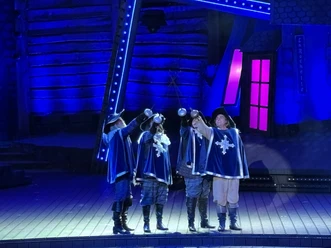 Freilichtbühne Meppen, 3 Musektiere - Das Musical 2024 ©Emsland Tourismus GmbH (2).jpeg Vier Musketiere in blauen Kostümen stoßen mit Schwertern auf der Freilichtbühne unter blauem Licht an.