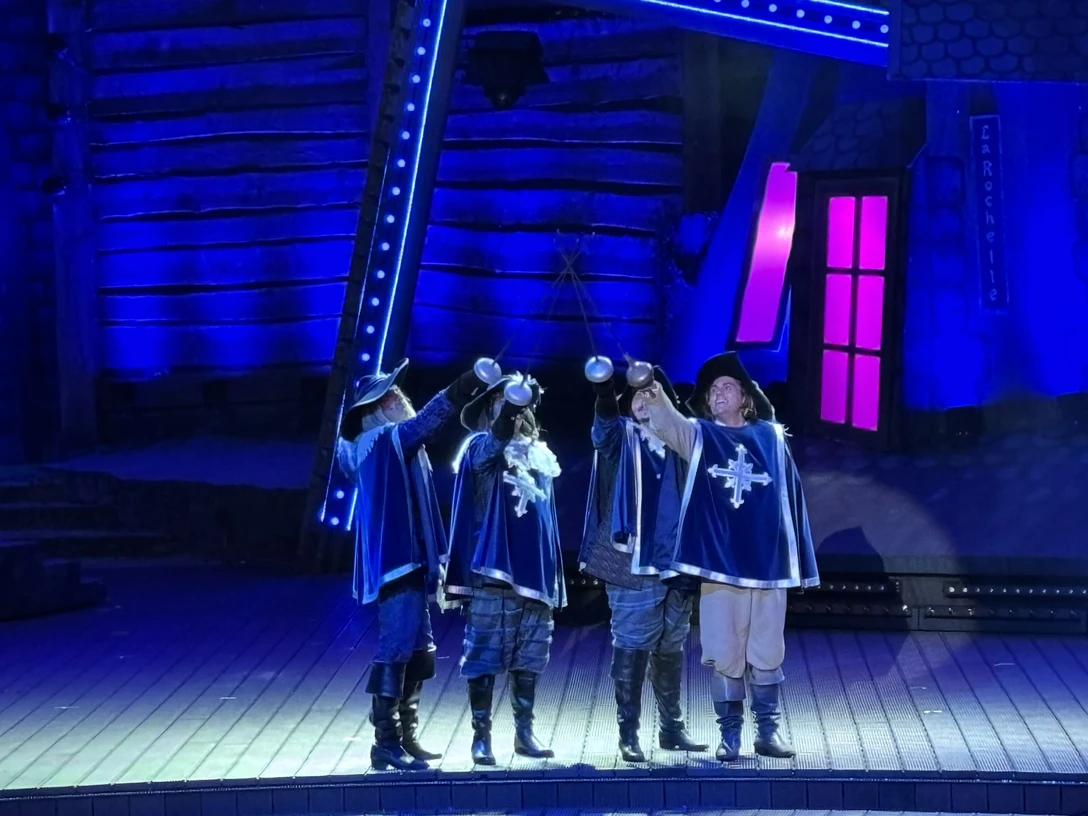 Freilichtbühne Meppen, 3 Musektiere - Das Musical 2024 ©Emsland Tourismus GmbH (2).jpeg Vier Musketiere in blauen Kostümen stoßen mit Schwertern auf der Freilichtbühne unter blauem Licht an.