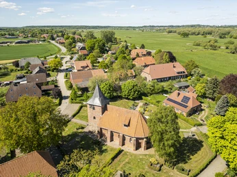 Die Kirche in Radegast aus der LuftperspektiveAerial view of Radegast village with a red-brick church surrounded by green fields and rural homesVue aérienne d’une église en briques entourée de maisons et de champs sous un ciel clair.Luchtfoto van de bakstenen kerk van Radegast te midden van rode daken en groen platteland.Kyrka i röd tegel med torn och spira, omgiven av gröna träd och lantliga hus i Radegast.Luftfoto af Radegast kirke omgivet af rødstenshuse, grønne marker og træer under blå himmel.