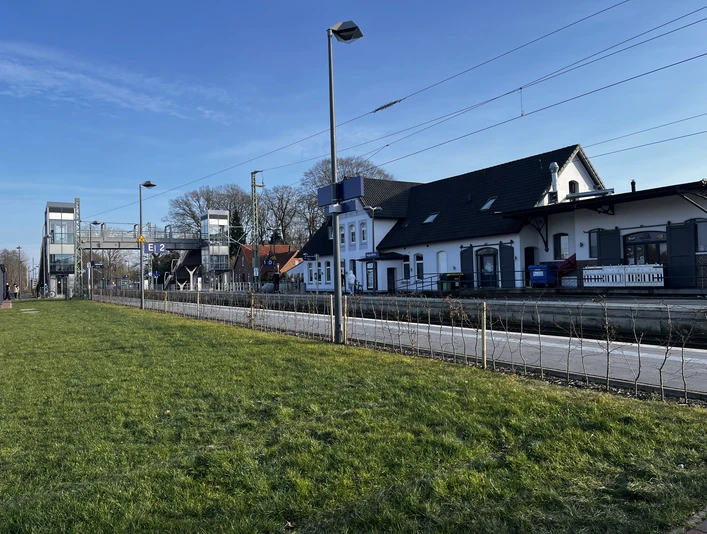 Bahnhof Augustfehn mit Fahrstuhl_2025 Bahnhof Augustfehn mit einem modernen Aufzug, Gleise, historische Gebäude und blauer Himmel im Hintergrund.