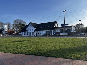 Augustfehn Bahnhof_Ansicht vom Dockgelände.jpg Der Bahnhof von Augustfehn zeigt ein saniertes, zweistöckiges Gebäude mit weißen Fassaden und dunklem, geneigtem Dach. Im Vordergrund erstreckt sich eine gepflegte Rasenfläche. Eisenbahngleise verlaufen entlang des Gebäudes, unter einem hellen, klaren Himmel.
