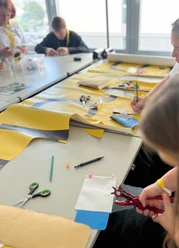re:create Workshop <p>Menschen basteln an einem Tisch mit buntem Papier und Werkzeugen in einer hellen Umgebung.</p>
