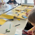 re:create Workshop <p>Menschen basteln an einem Tisch mit buntem Papier und Werkzeugen in einer hellen Umgebung.</p>