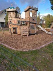 Holzspielplatz mit Klettergerüst, Rutsche und Kletterwand, umgeben von Holzhackschnitzeln und Bäumen.