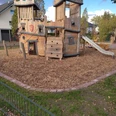 Holzspielplatz mit Klettergerüst, Rutsche und Kletterwand, umgeben von Holzhackschnitzeln und Bäumen.