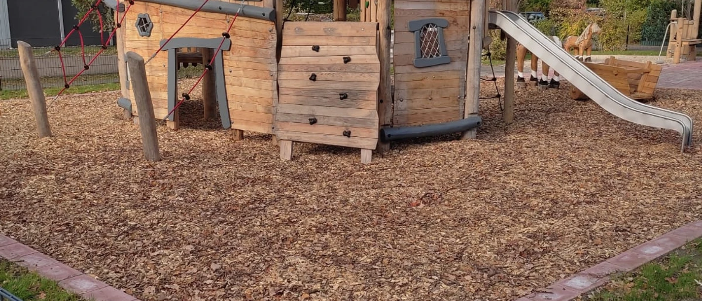 Spielplatz Alleenviertel Holzspielplatz mit Klettergerüst, Rutsche und Kletterwand, umgeben von Holzhackschnitzeln und Bäumen.