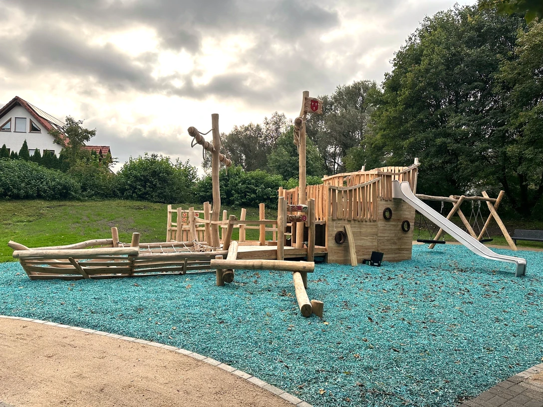 Bild_Spielplatz am Dedinger-Heide-See.PNG Spielplatz mit Piratenschiff-Thema aus Holz, umgeben von Bäumen, mit Rutsche und Kletterelementen.