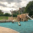 Bild_Spielplatz am Dedinger-Heide-See.PNG Spielplatz mit Piratenschiff-Thema aus Holz, umgeben von Bäumen, mit Rutsche und Kletterelementen.