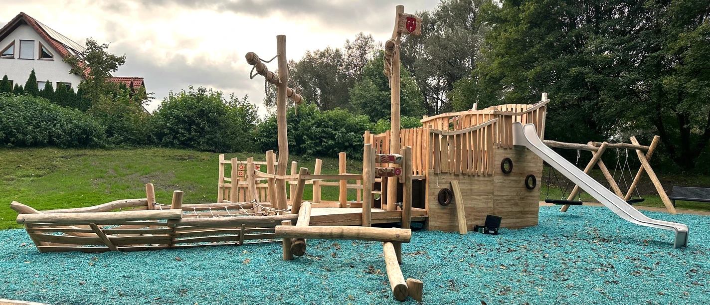 Bild_Spielplatz am Dedinger-Heide-See.PNG Spielplatz mit Piratenschiff-Thema aus Holz, umgeben von Bäumen, mit Rutsche und Kletterelementen.