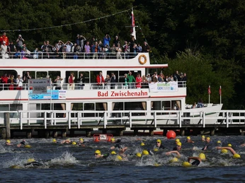 quer-durchs-meer Schwimmer mit gelben Badekappen im Wettkampf, begleitet von einem Ausflugsboot voller Zuschauer.