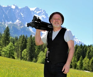 Jo Jonietz Mann mit Fotostativ auf Schulter, lächelnd vor Bergkulisse mit Wiesen und Wald.