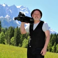 Jo Jonietz Mann mit Fotostativ auf Schulter, lächelnd vor Bergkulisse mit Wiesen und Wald.