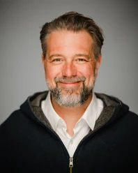 Fotograf Max Merget Ein lächelnder Mann mit Bart und grauen Haaren vor grauem Hintergrund.