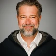 Fotograf Max Merget Ein lächelnder Mann mit Bart und grauen Haaren vor grauem Hintergrund.