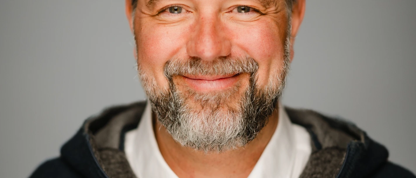 Fotograf Max Merget Ein lächelnder Mann mit Bart und grauen Haaren vor grauem Hintergrund.