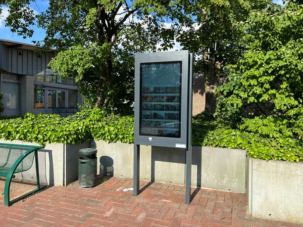 A modern information kiosk stands in a paved plaza, surrounded by trees and bushes.