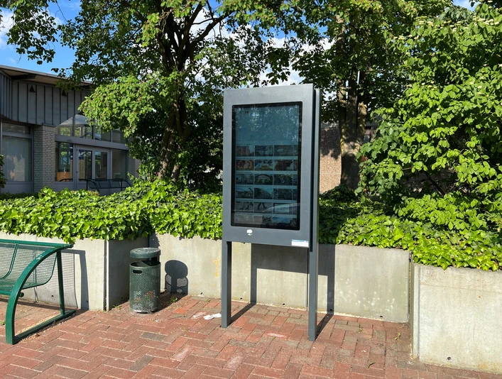 Infoterminal Eystrup Ein modernes Infoterminal steht auf einem gepflasterten Platz, umgeben von Bäumen und Büschen.