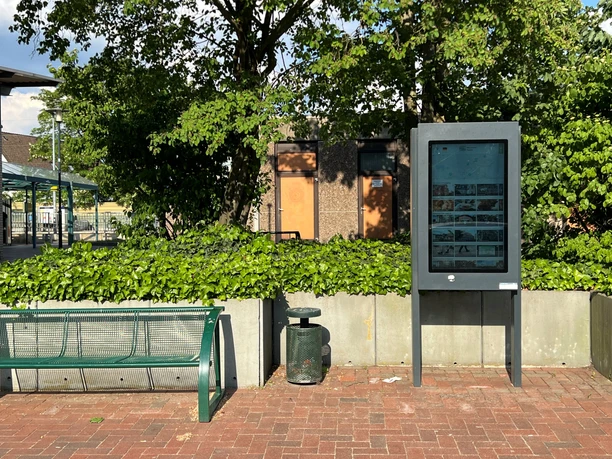A modern information kiosk stands in front of green bushes on a paved surface in a sunny setting.