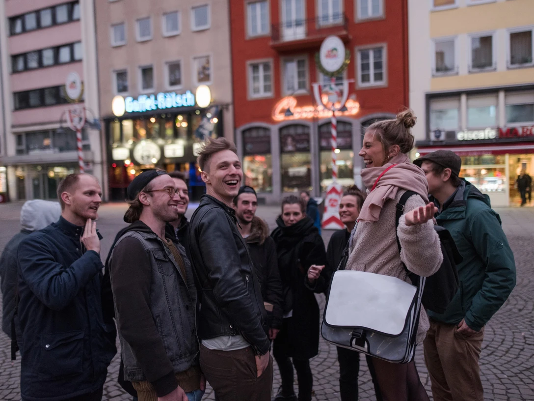 Eine fröhliche Gruppe erkundet lächelnd die Altstadt von Köln, umgeben von historischen Gebäuden und typisch kölschen Brauhäusern.A cheerful group smiles as they explore Cologne's old town, surrounded by historic buildings and typical Cologne breweries.