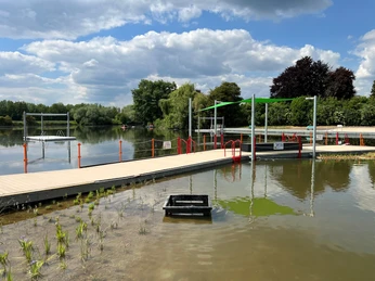 Naturbad Landesbergen Naturbad mit Steg und Zugangshilfen an einem ruhigen See, umgeben von Bäumen und bewölktem Himmel.