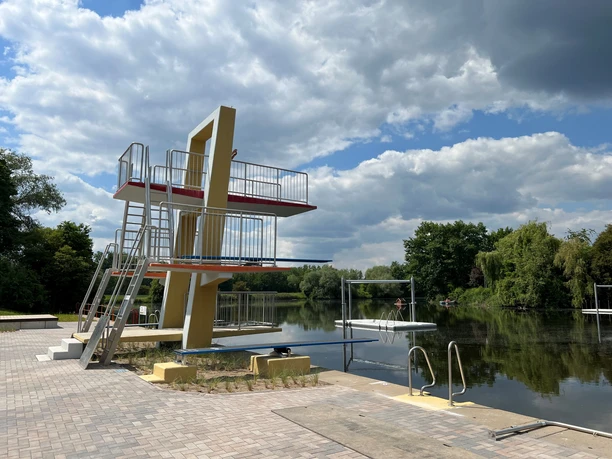 Naturbad Landesbergen Ein modernes Naturbad mit einem Sprungturm aus Metall und Beton am ruhigen See an einem bewölkten Tag.