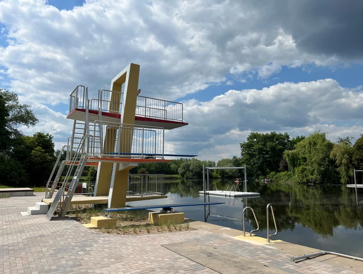 Naturbad Landesbergen Ein modernes Naturbad mit einem Sprungturm aus Metall und Beton am ruhigen See an einem bewölkten Tag.