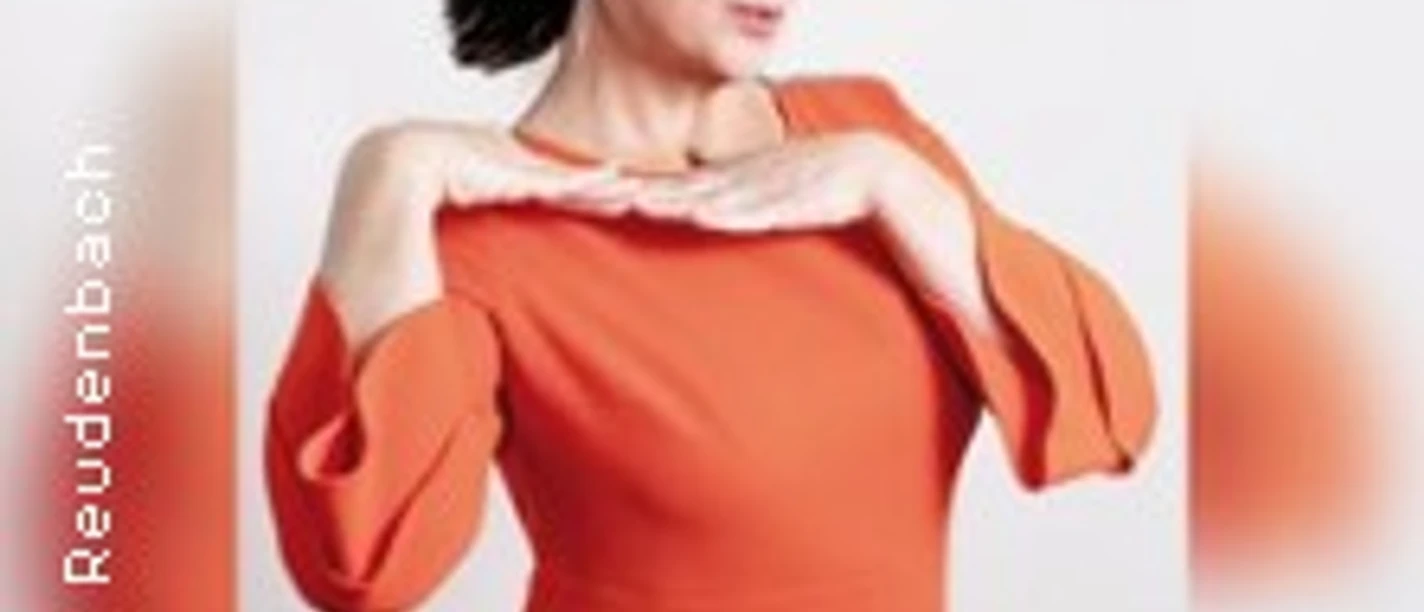 Frau in orangefarbenem Kleid posiert selbstbewusst mit Händen am Kinn vor weißem Hintergrund.