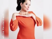 Frau in orangefarbenem Kleid posiert selbstbewusst mit Händen am Kinn vor weißem Hintergrund.