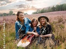 Drei Personen in legerer Kleidung sitzen lächelnd in einer blühenden Heidelandschaft bei Sonnenuntergang.