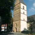 kirche.jpg Ein historischer Kirchturm aus Sandstein, umgeben von Bäumen, vor einem blauem Himmel.