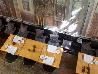 Christoph Pauls Restaurant Im Innenraum von Christoph Pauls Restaurant erstreckt sich ein beeindruckendes Wandgemälde, das mit gotischen Spitzbögen und bunten Glasfenstern dekoriert ist. Darunter stehen adrett gedeckte Holztische, die eine einladende Atmosphäre schaffen.Inside Christoph Paul's restaurant extends an impressive mural, decorated with Gothic pointed arches and colorful stained glass windows. Neatly set wooden tables stand beneath, creating a welcoming atmosphere.