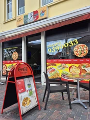 Pizzaria La Rosa Die Außenansicht der Pizzeria "La Rosa" zeigt ein gelbes Gebäude mit markantem roten Schild und großen Fenstern, die bunte Werbeaufkleber für verschiedene Gerichte tragen. Vor der Eingangstür stehen zwei schwarze Stühle und ein rotes Plakat für das Tagesangebot.