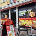 Pizzaria La Rosa Die Außenansicht der Pizzeria "La Rosa" zeigt ein gelbes Gebäude mit markantem roten Schild und großen Fenstern, die bunte Werbeaufkleber für verschiedene Gerichte tragen. Vor der Eingangstür stehen zwei schwarze Stühle und ein rotes Plakat für das Tagesangebot.