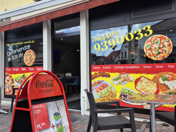 Pizzaria La Rosa Die Außenansicht der Pizzeria "La Rosa" zeigt ein gelbes Gebäude mit markantem roten Schild und großen Fenstern, die bunte Werbeaufkleber für verschiedene Gerichte tragen. Vor der Eingangstür stehen zwei schwarze Stühle und ein rotes Plakat für das Tagesangebot.