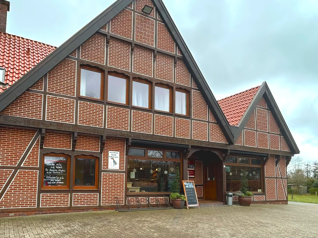 Landschlachterei Ammerland Aussenansicht Das Bild zeigt ein traditionelles, norddeutsches Backsteingebäude mit rotem Ziegeldach und einer Fachwerkstruktur. Die Fassade des Gebäudes ist von mehreren Holzunterteilungen durchzogen, und Fenster sowie Eingänge sind symmetrisch angeordnet. Der Himmel ist bewölkt, was der Szene eine ruhige Atmosphäre verleiht.