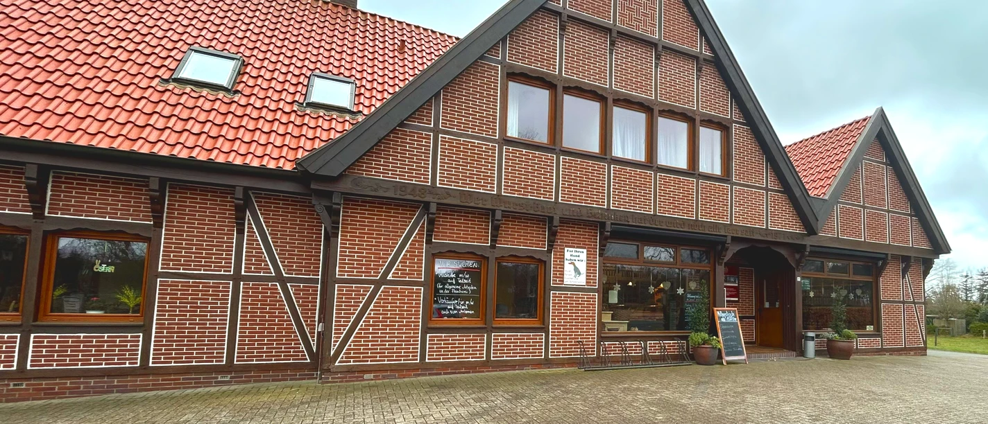 Landschlachterei Ammerland Aussenansicht Das Bild zeigt ein traditionelles, norddeutsches Backsteingebäude mit rotem Ziegeldach und einer Fachwerkstruktur. Die Fassade des Gebäudes ist von mehreren Holzunterteilungen durchzogen, und Fenster sowie Eingänge sind symmetrisch angeordnet. Der Himmel ist bewölkt, was der Szene eine ruhige Atmosphäre verleiht.