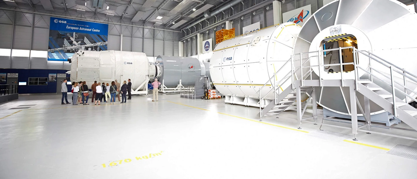 Deutsches-Zentrum-fuer-Luft-und-Raumfahrt-Gruppe-Axel-Schulten-KoelnTourismus-GmbH.jpg Innenansicht des Deutschen Zentrums für Luft- und Raumfahrt. Im Vordergrund ist eine Gruppe von Personen, die eine Führung durch eine große Halle erhalten, in der verschiedene Raumfahrmodule und technische Einrichtungen zu sehen sind.