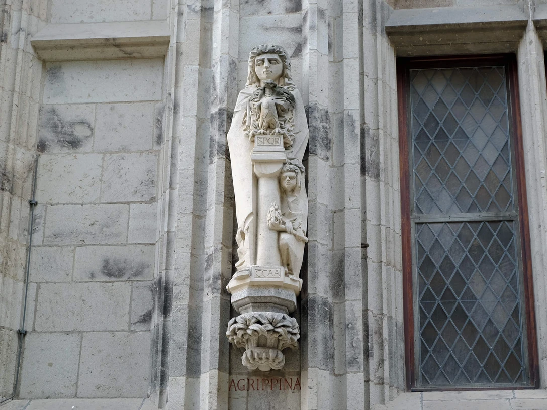 Agrippina-Statue am Rathausturm in Köln: Steinskulptur einer Frau mit Kranz neben verzierten Fenstern.Agrippina statue on the town hall tower in Cologne: stone sculpture of a woman with a wreath next to decorated windows.