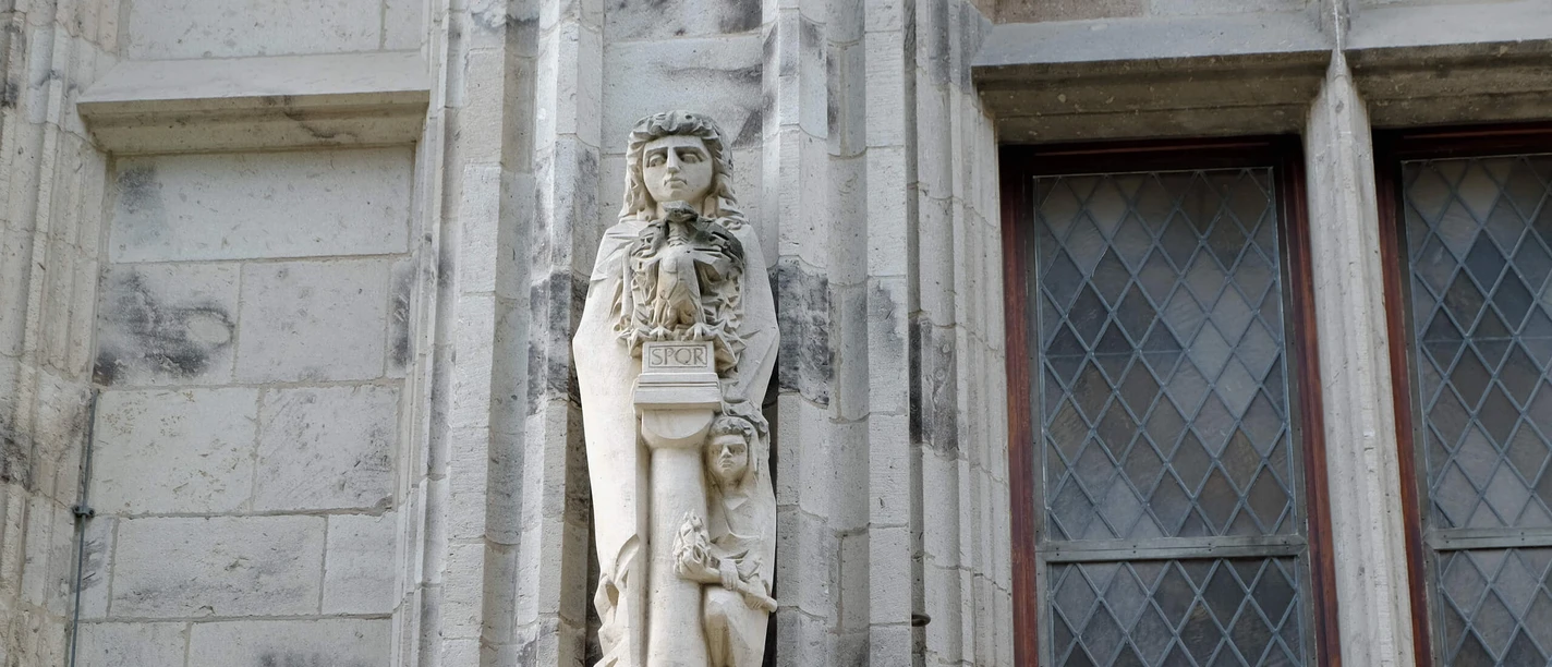 Agrippina statue on the town hall tower in Cologne: stone sculpture of a woman with a wreath next to decorated windows.