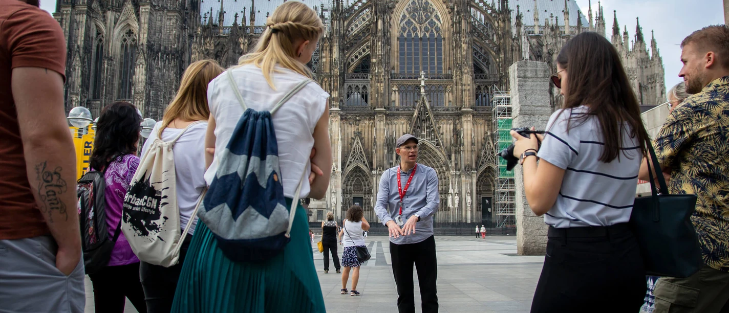 A group of tourists listen to a city guide on Roncalliplatz, with the majestic Cologne Cathedral in the background. The sky is slightly overcast, giving the scene a lively yet relaxed atmosphere.