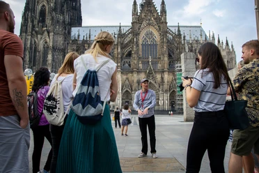 Stadtfuehrung-Roncalliplatz-Bilderblitz-KoelnTourismus-GmbH.jpg Eine Gruppe von Tourist*innen lauscht einem Stadtführer auf dem Roncalliplatz, mit dem majestätischen Kölner Dom im Hintergrund. Der Himmel ist leicht bewölkt, was der Szene eine lebendige und zugleich entspannte Atmosphäre verleiht.