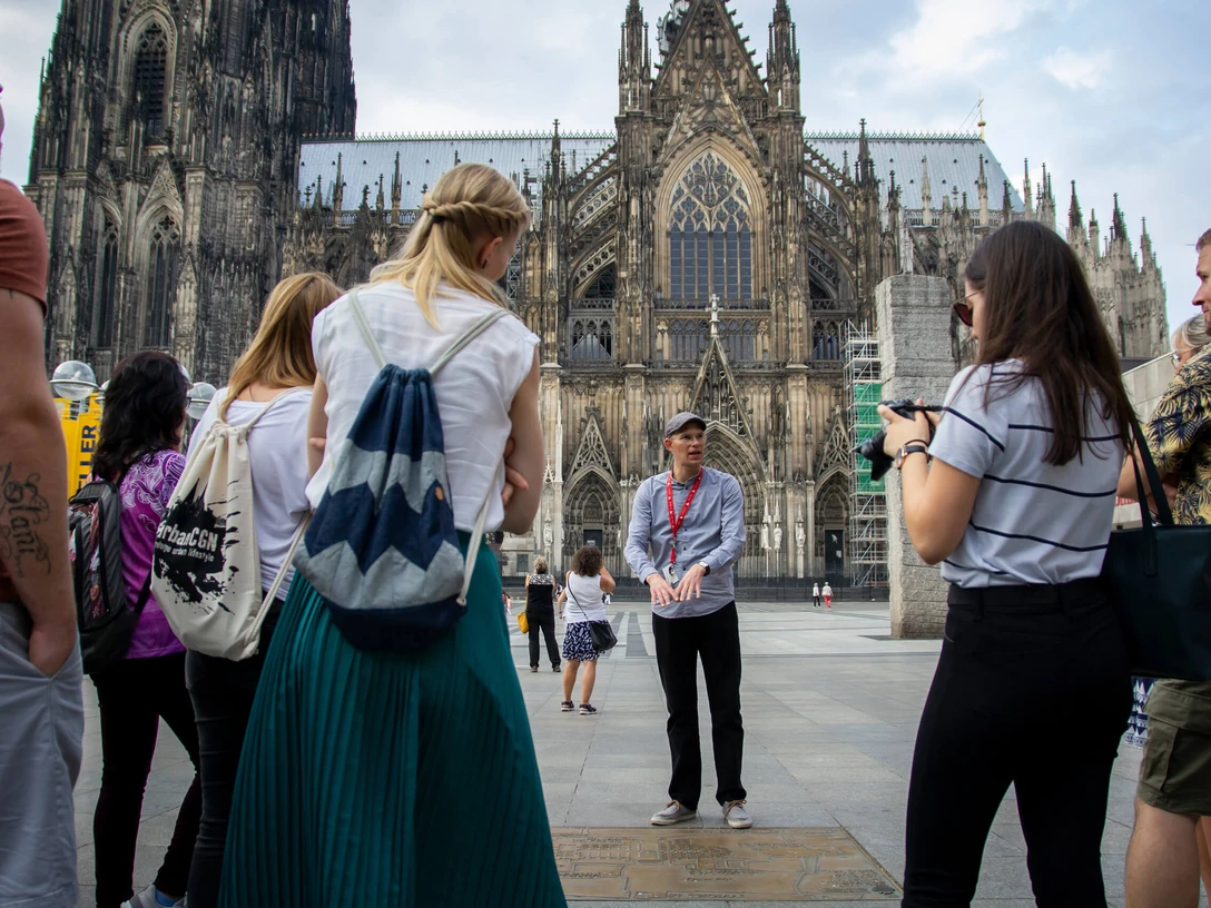 Stadtfuehrung-Roncalliplatz-Bilderblitz-KoelnTourismus-GmbH.jpg Eine Gruppe von Tourist*innen lauscht einem Stadtführer auf dem Roncalliplatz, mit dem majestätischen Kölner Dom im Hintergrund. Der Himmel ist leicht bewölkt, was der Szene eine lebendige und zugleich entspannte Atmosphäre verleiht.A group of tourists listen to a city guide on Roncalliplatz, with the majestic Cologne Cathedral in the background. The sky is slightly overcast, giving the scene a lively yet relaxed atmosphere.
