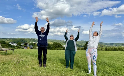 Wanderung mit Atem- und Yogaübungen Drei Personen stehen auf einer Wiese und strecken ihre Arme fröhlich zum strahlend blauen Himmel.