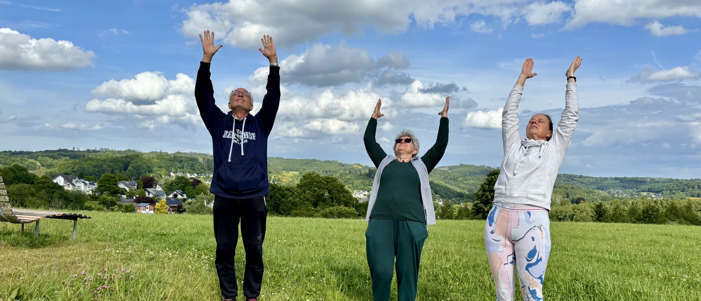 Wanderung mit Atem- und Yogaübungen Drei Personen stehen auf einer Wiese und strecken ihre Arme fröhlich zum strahlend blauen Himmel.