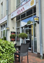Restaurant Kibar Vor einem modernen Gebäude mit hohen Glasfenstern steht das Restaurant Kibar. Zwei Topfpflanzen flankieren den Eingang, daneben befinden sich draußen Tische und Stühle für Gäste.
