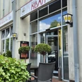 Restaurant Kibar Vor einem modernen Gebäude mit hohen Glasfenstern steht das Restaurant Kibar. Zwei Topfpflanzen flankieren den Eingang, daneben befinden sich draußen Tische und Stühle für Gäste.