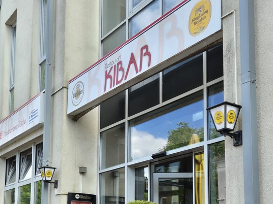 Restaurant Kibar Vor einem modernen Gebäude mit hohen Glasfenstern steht das Restaurant Kibar. Zwei Topfpflanzen flankieren den Eingang, daneben befinden sich draußen Tische und Stühle für Gäste.