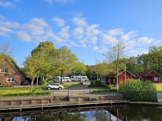 240429_Wohnmobilstellplatz-Uferstraße_Uplengen.jpg Vier Wohnmobile im Hintergrund, im Vordergrund rote Hütten mit einem Steg und Wasser, Kanal