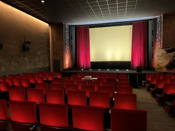 Filmhof Hoya Ein leerer Kinosaal mit roten Sitzen und einem beleuchteten Bildschirm, umrahmt von roten Vorhängen.