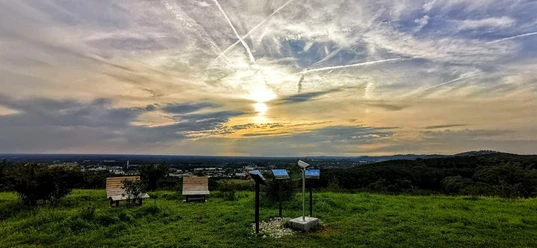 panorama_grosser_berg.jpg Sonnenuntergang über einer grünen Landschaft mit Info-Tafeln und Parkbänken unter dramatischem Himmel.